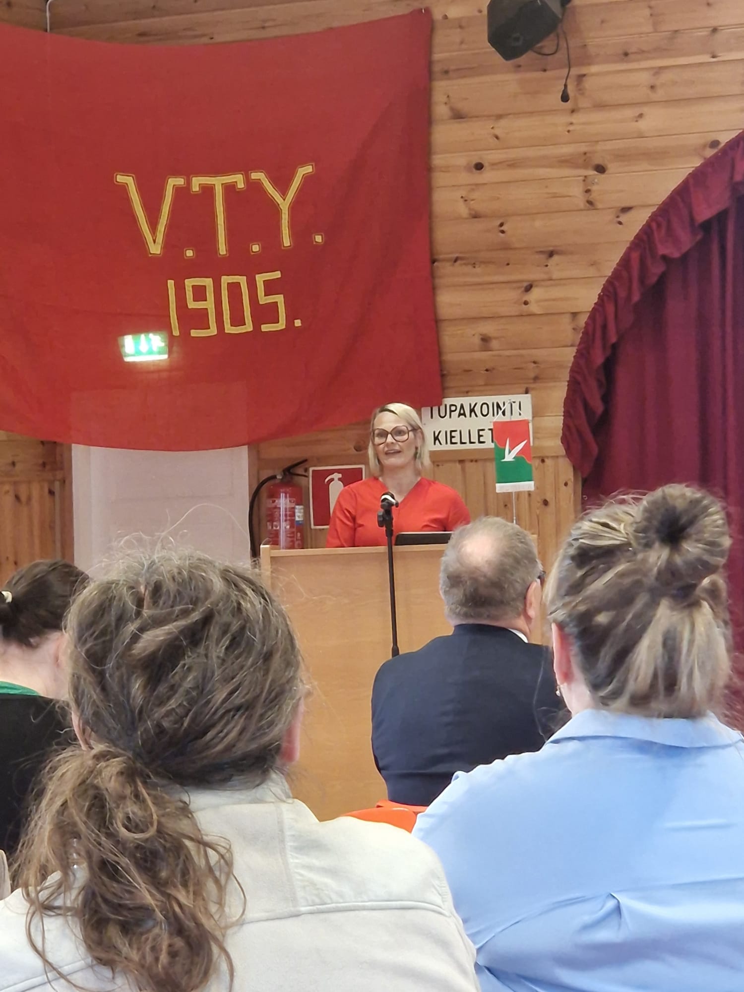Vaaleahiuksinen keski-ikäinen nainen, jolla on punasankaiset isot silmälasit, seisoo puhujanpöydän takana puhetta pitämässä. Taustalla iso punainen lakana, jossa lukee V.T.Y. 1905. Kuva on otettu yleisön seasta kauempaa, ja edessä näkyy neljän kuulija takaraivot. 